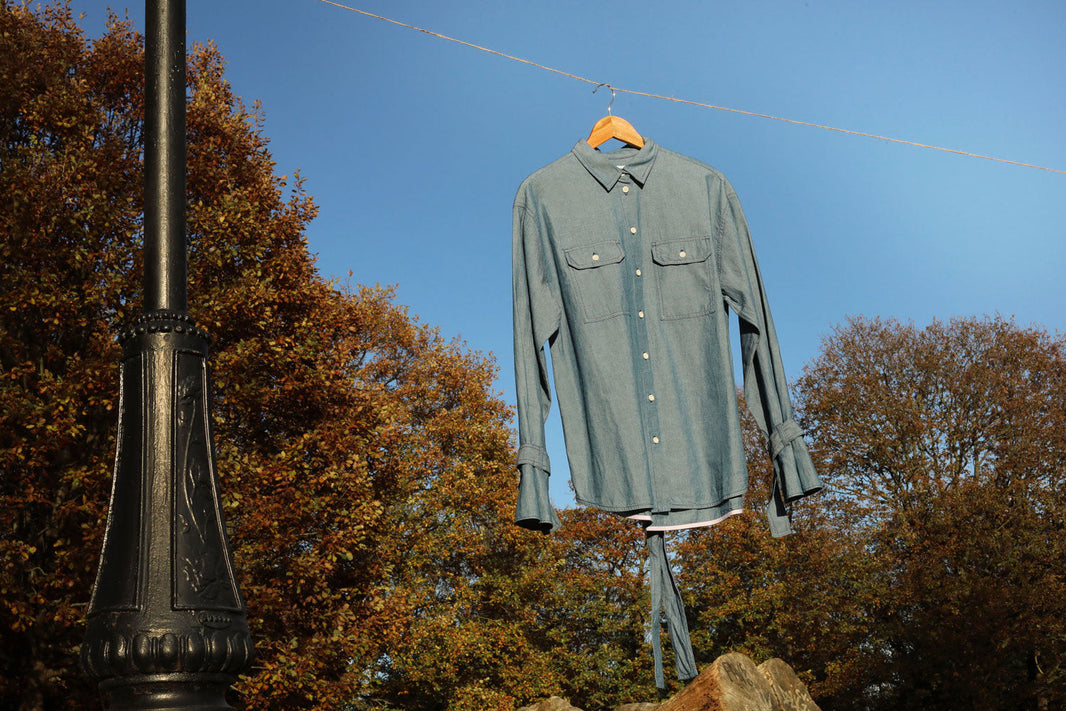 Women's Japanese Denim Boyfriend Shirt by Saywood, the Zadie Lightwash Denim Shirt, hanging on a hanger on a line of natural string, swinging in the breeze. A black lamp post is seen to the left of the image with trees and blue sky in the background.