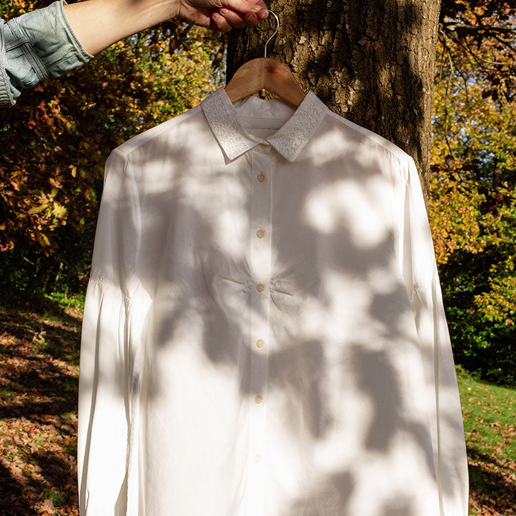 Saywood's Edi White Shirt with lace collar and volume sleeves, made in London, is hanging on a wooden hanger, being held by a hand with a denim shirt sleeve just visible. The shirt is hanging in front of a tree with Autumnal leafy tress in the background.