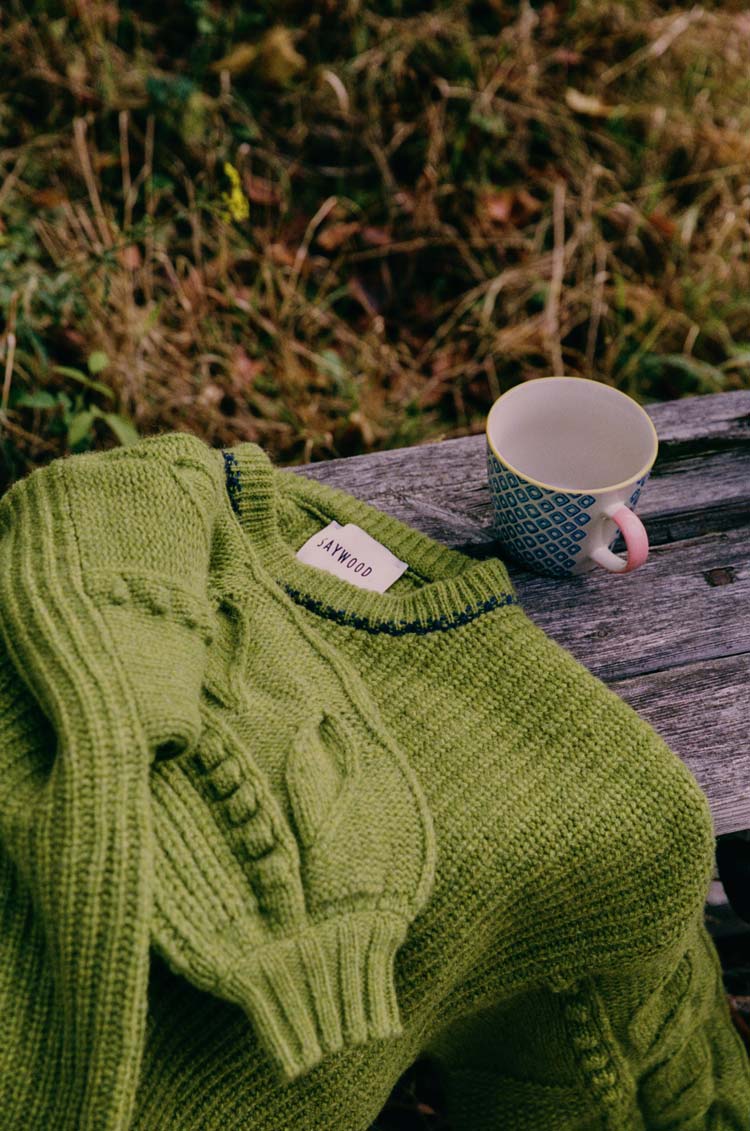 Saywood moss green lambswool fisherman rib knit jumper folded on a weathered wooden bench beside a ceramic mug — British made knitwear, crafted to be cared for and kept