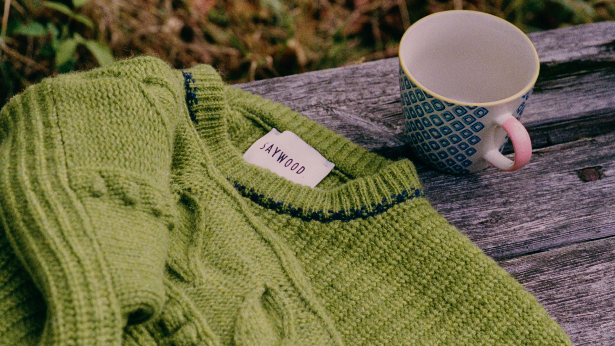Saywood moss green lambswool fisherman rib knit jumper folded on a weathered wooden bench beside a ceramic mug — British made knitwear, crafted to be cared for and kept