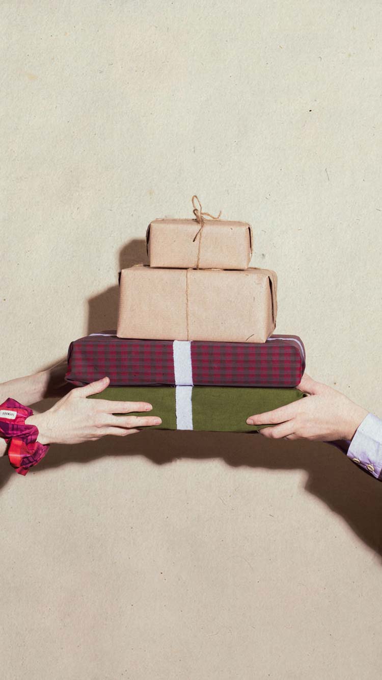 Two pairs of hands exchanging a stack of beautifully wrapped Christmas gifts — kraft paper, tartan and forest green — sustainable luxury gifting by Saywood