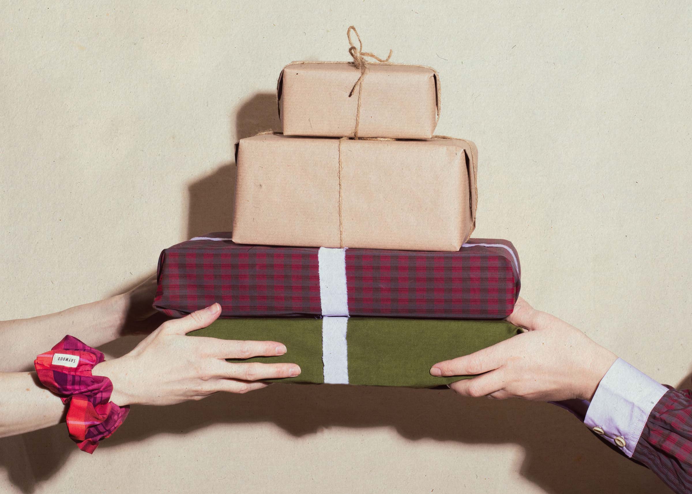 Two pairs of hands exchanging a stack of beautifully wrapped Christmas gifts — kraft paper, tartan and forest green — sustainable luxury gifting by Saywood