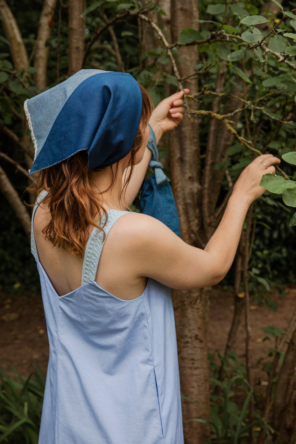 Maya Cami Dress in pale blue recycled cotton, back view with tie belt and headscarf, model reaching up among trees. By Saywood. Made in London.