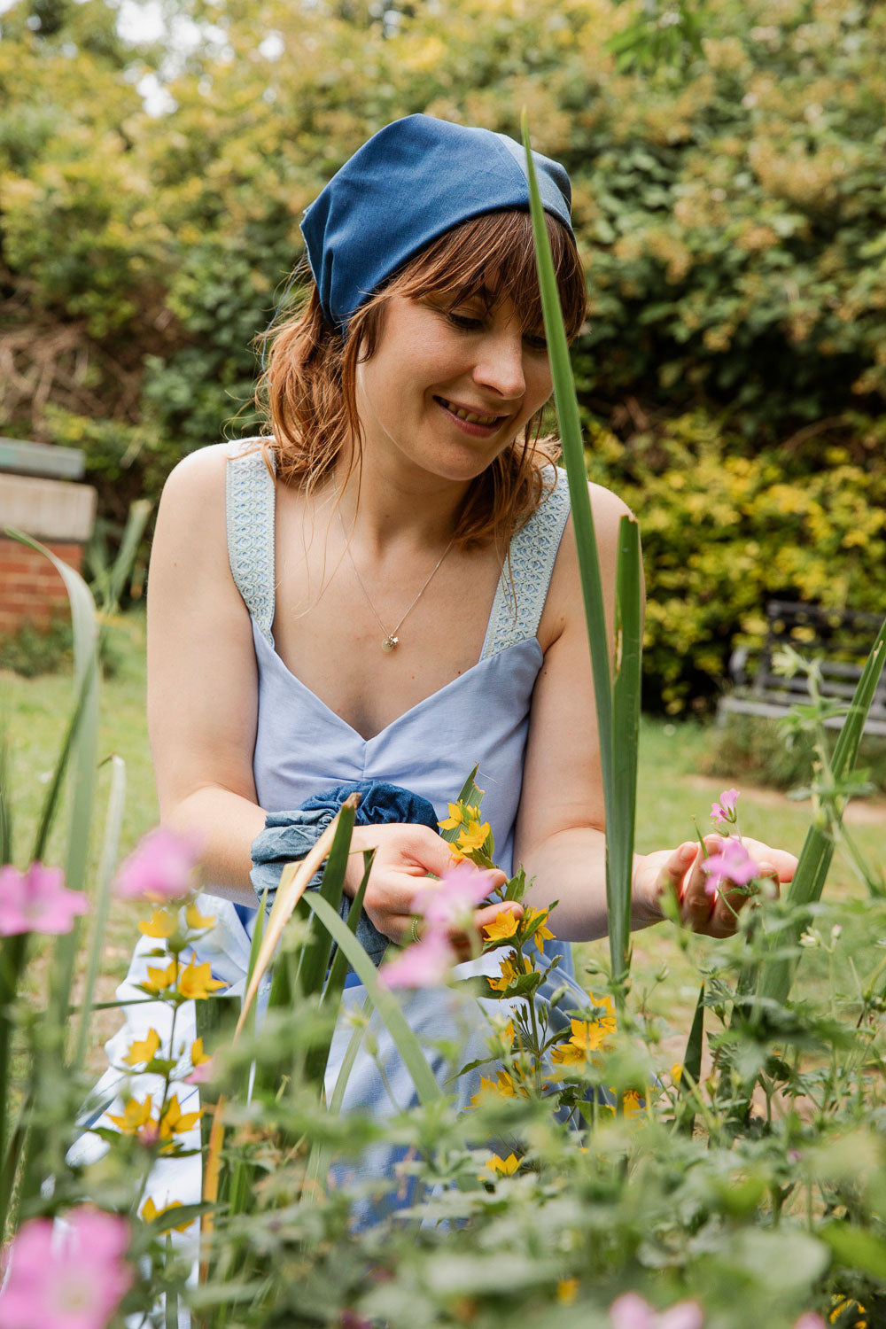 Maya Cami Dress in pale blue recycled cotton, worn with detachable tie belt, model crouching among wildflowers. By Saywood. Made in London.