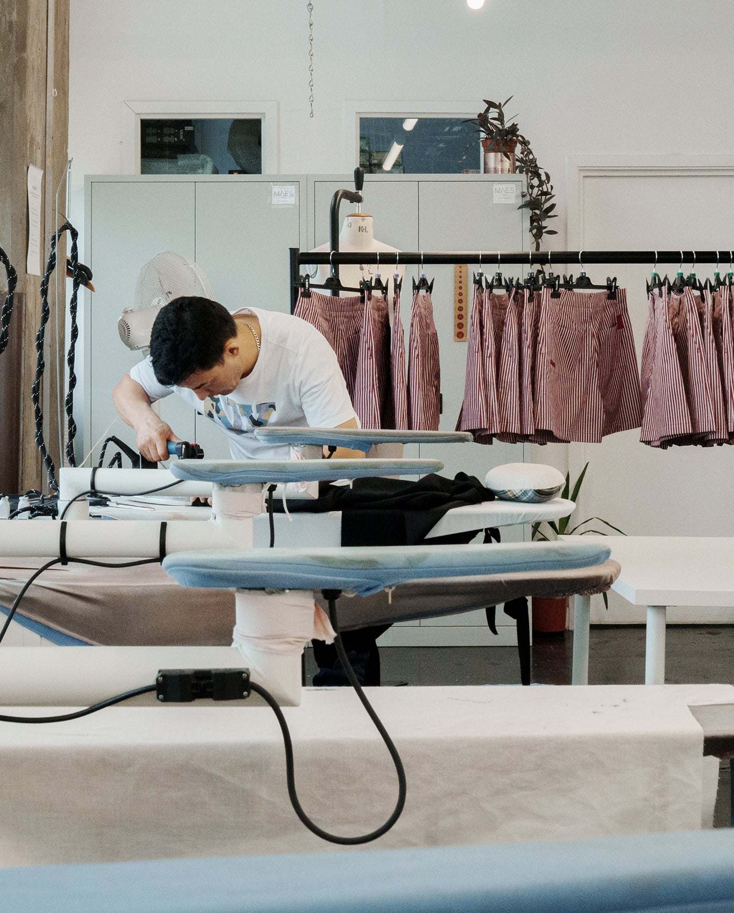 A man is pressing garments on an ironing board in a factory. Saywood's red and white stripe Bessie A-line Shorts are on a rail behind him.