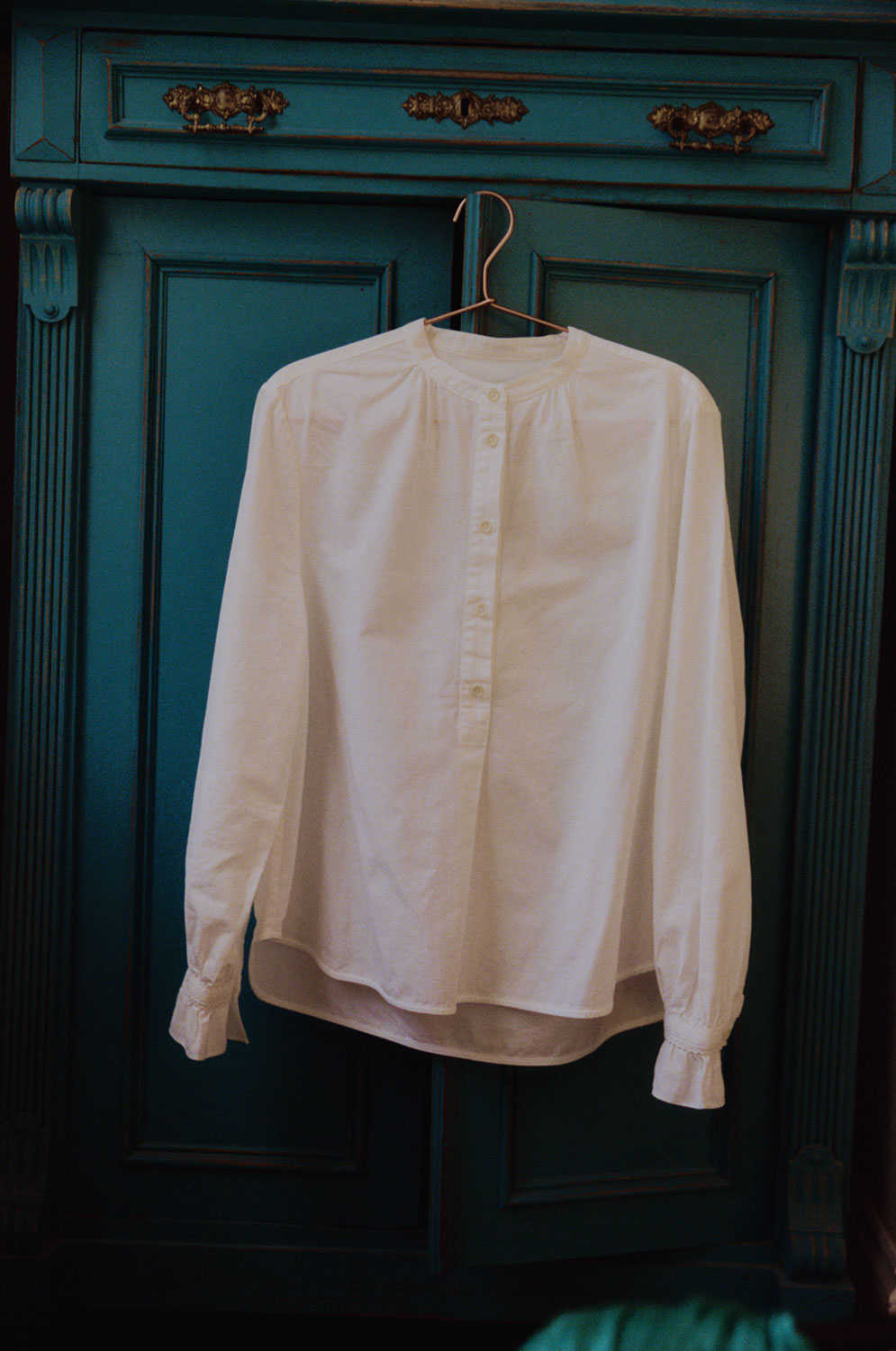 Marie white A-line blouse in recycled cotton with gathered neck and lace-trimmed cuffs by Saywood, hanging on wardrobe.