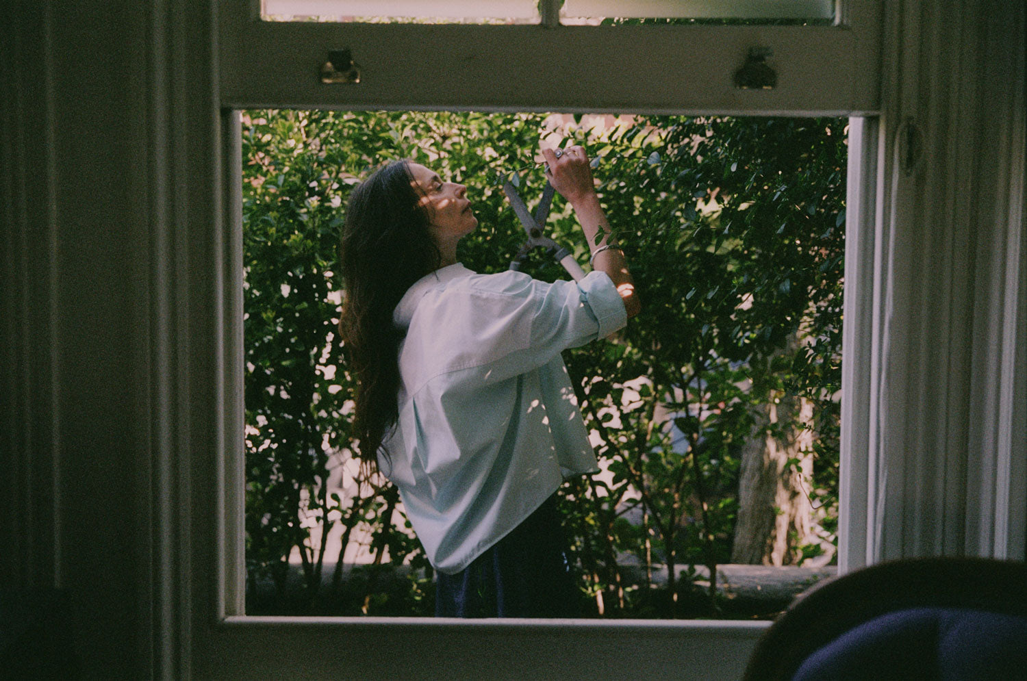 A women is standing outside, with the camera shooting through an open sash window. She is wearing Saywood's Lela Colourblock Boxy Shirt in pastel green and blue, with the sleeve rolled up, and the Japanese Denim Amelia Trousers. She is holding a large pair of hedge cutters and reaching out to the leafy hedge.