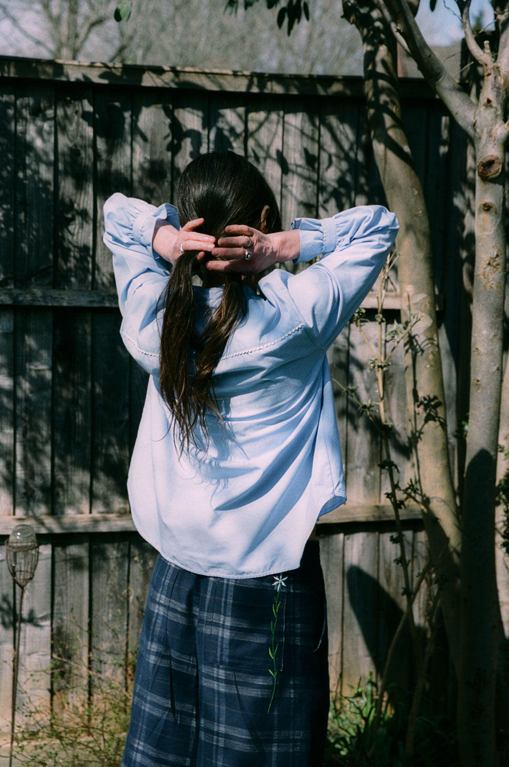 Back view, Marie pale blue lace-trimmed blouse with gathered cuffs, A-line silhouette, made in London by Saywood. Worn with navy check trousers.