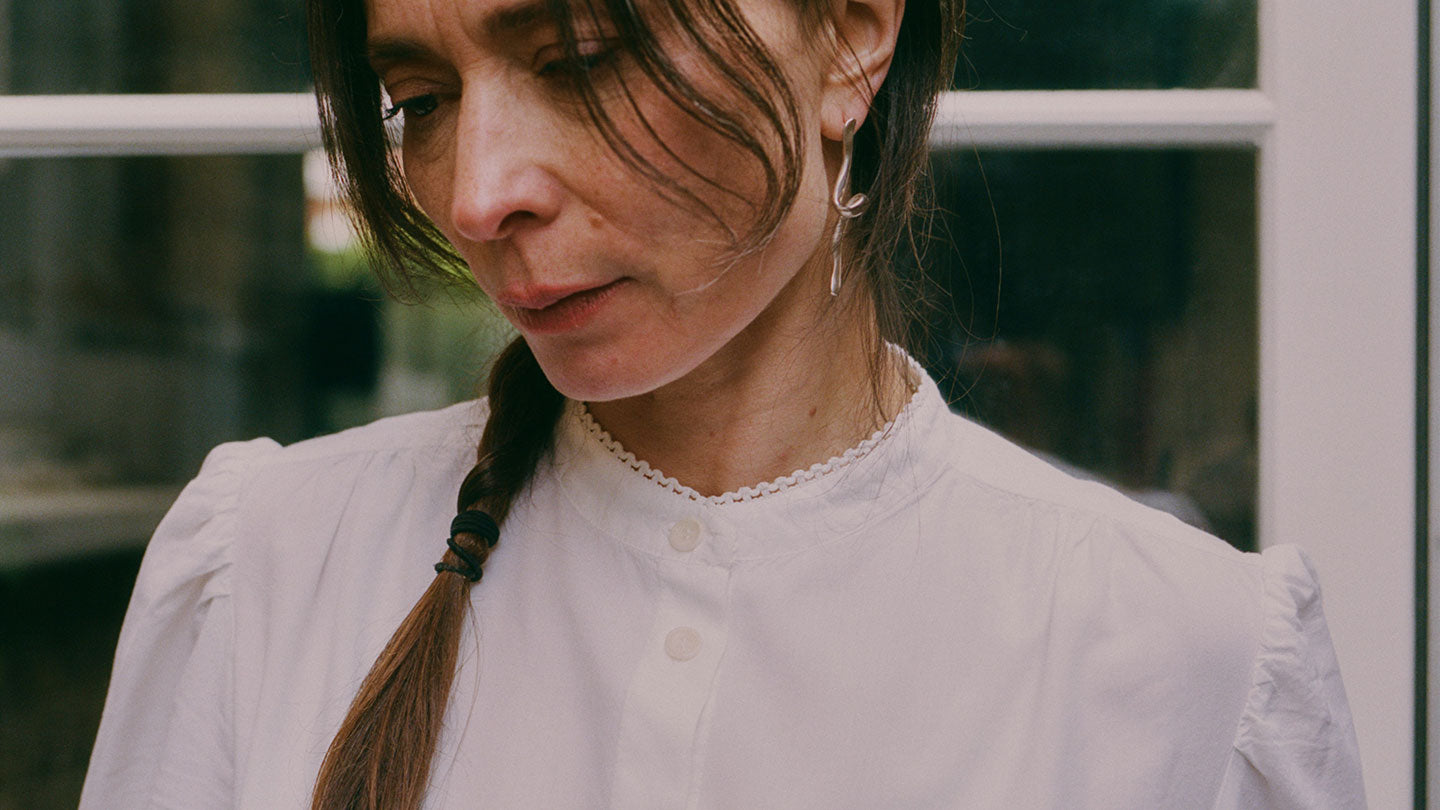 Close up of a woman wearing Saywood's white Joni Puff sleeve Blouse, with lace trim around the edge of the grandad collar. She wears her hair in a plait, with a silver twisted drop earring in her ear. She is standing in front of a white glass paned door.