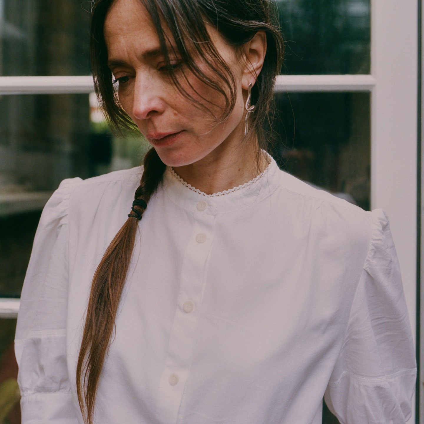 Close up of a woman wearing Saywood's white Joni Puff sleeve Blouse, with lace trim around the edge of the grandad collar. She wears her hair in a plait, with a silver twisted drop earring in her ear. She is standing in front of a white glass paned door.